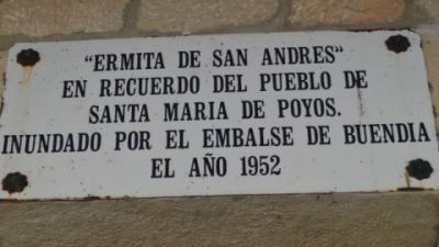 Una placa blanca rectangular, desgastada por el tiempo y fijada a una pared de piedra, conmemora la ermita de San Andrés y el pueblo de Santa María de Poyos, inundado en 1952 por el embalse de Buendía.  La placa tiene texto en negro, con letras mayúsculas y puntuación clara.