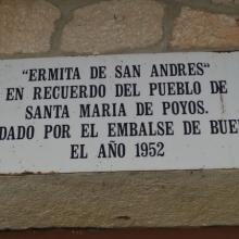 Una placa blanca rectangular, desgastada por el tiempo y fijada a una pared de piedra, conmemora la ermita de San Andrés y el pueblo de Santa María de Poyos, inundado en 1952 por el embalse de Buendía.  La placa tiene texto en negro, con letras mayúsculas y puntuación clara.