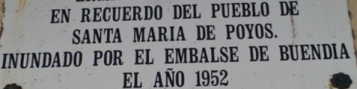 Una placa blanca rectangular, desgastada por el tiempo y fijada a una pared de piedra, conmemora la ermita de San Andrés y el pueblo de Santa María de Poyos, inundado en 1952 por el embalse de Buendía.  La placa tiene texto en negro, con letras mayúsculas y puntuación clara.